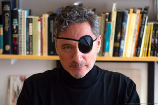 Headshot of Höcek (white man wearing a black eye patch, short silver hair) with bookshelf in the background.