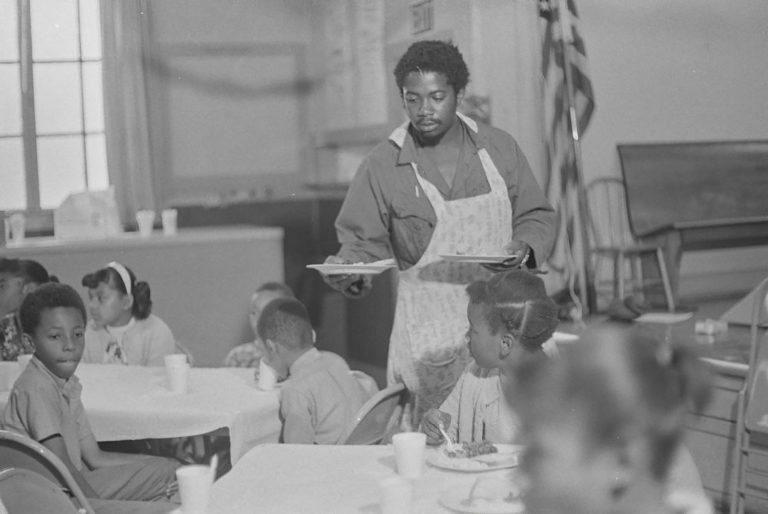 Black-and-white photograph of a dark-skinned man in an apron serving food to children sitting on tables
