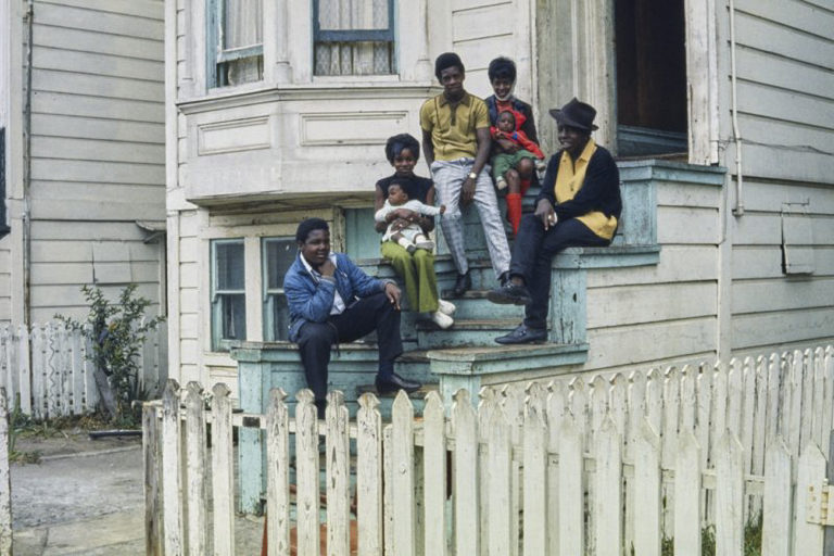 Three dark-skinned men, two dark-skinned women and two dark-skinned babies sit on the stoop of a white clapboard building