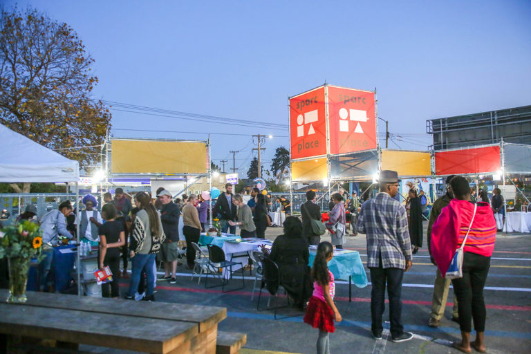 A large group of people gathered at dusk mill about tents and kiosks made of scaffolding with orange and yellow signage