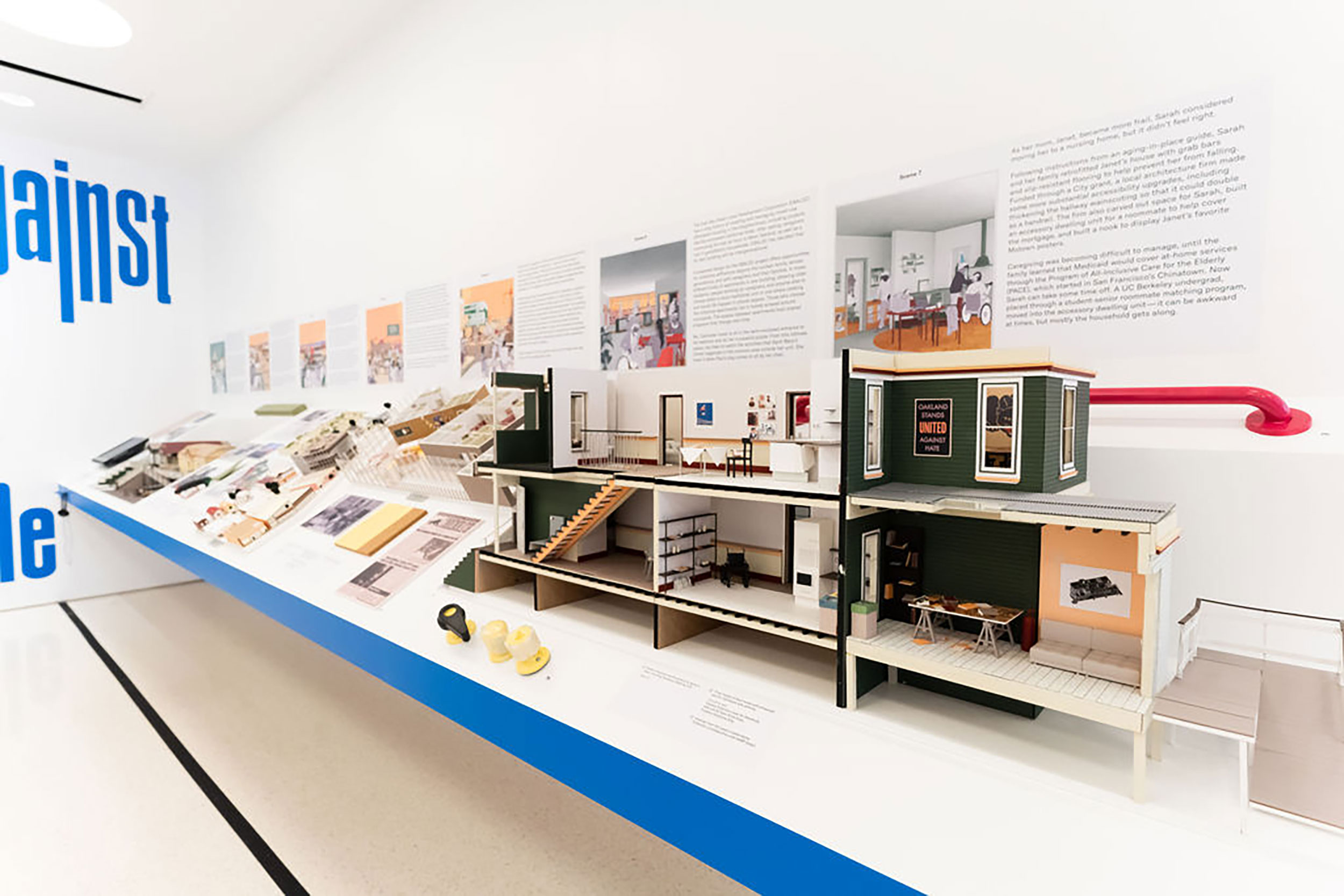 Installation view of a gallery wall with text and images, with a shelf that has 3D architectural models on it.
