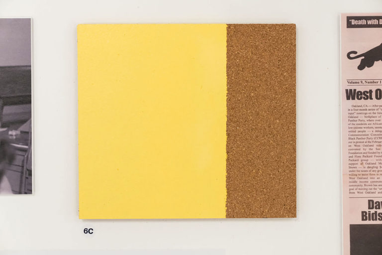 A square tile made out of cork, two thirds of which have been painted a glossy yellow color and the final third left bare.