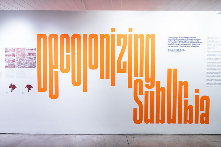 Installation view of a white gallery wall with large orange text and smaller black text.
