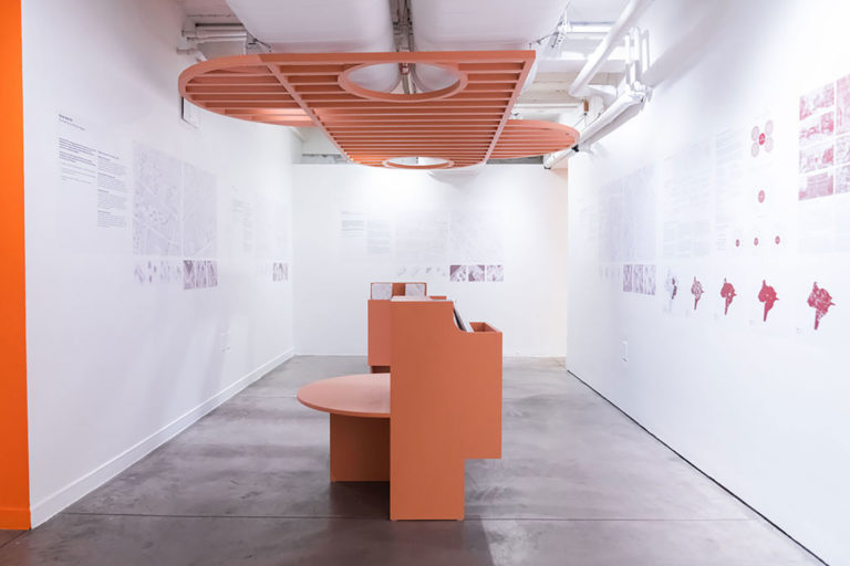 Installation view of a gallery room of white walls with text, a salmon-colored bench in the middle of the room, and a salmon-colored shaded roof above the bench attached to the ceiling.