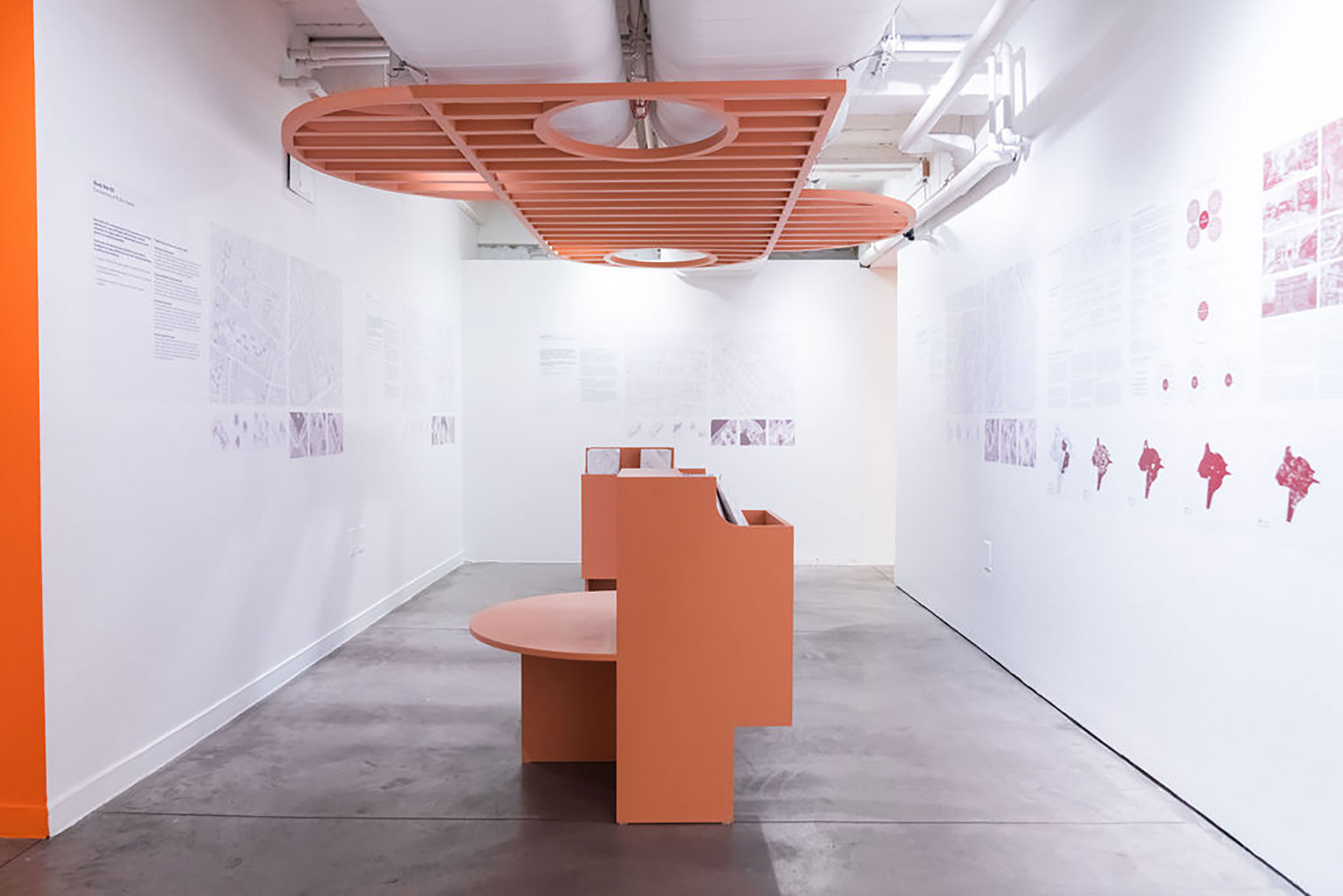 Installation view of a gallery room of white walls with text, a salmon-colored bench in the middle of the room, and a salmon-colored shaded roof above the bench attached to the ceiling.