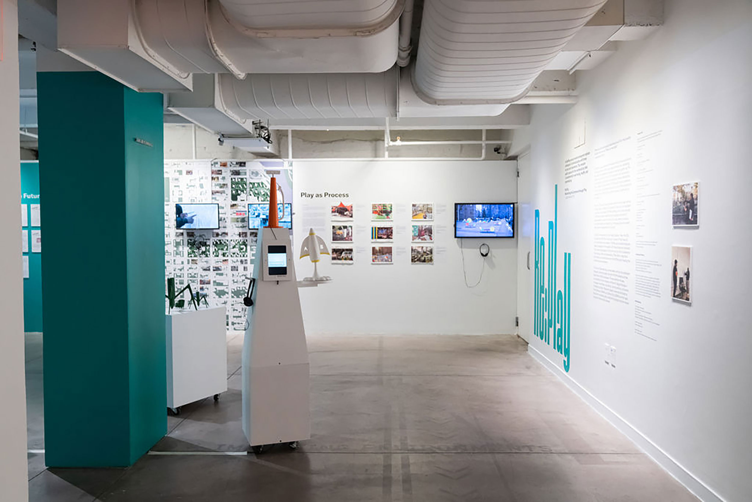 Installation view of a gallery room with white walls lined with text and TVS, blue details, and a free standing structure in the middle.