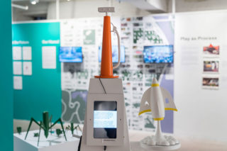 Installation view of gallery room focused on a white structure with orange top that holds a screen and a model of a rocket taking off.