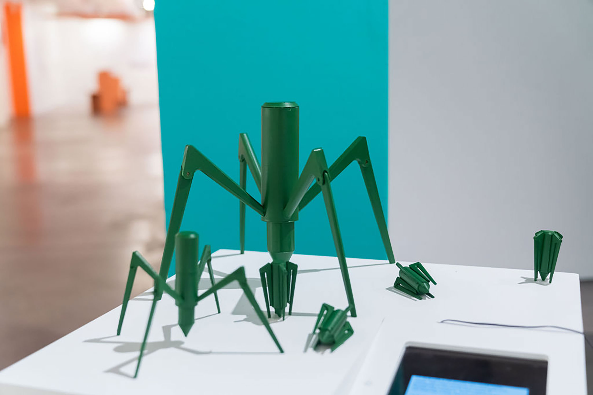 Installation view of small green objects on a white surface against a blue and white wall.