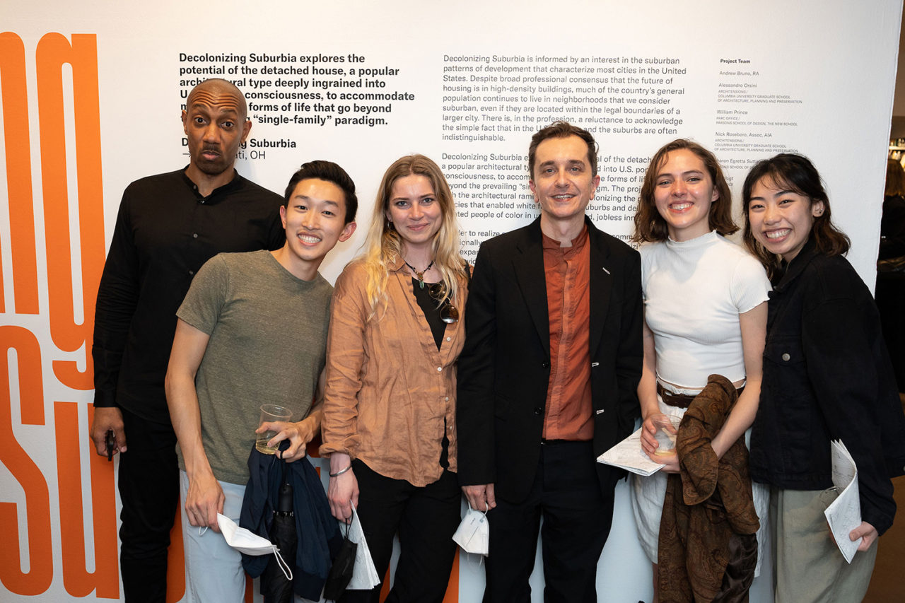 Six individuals stand in front of a white wall with large-scale graphic orange text and smaller text in black.