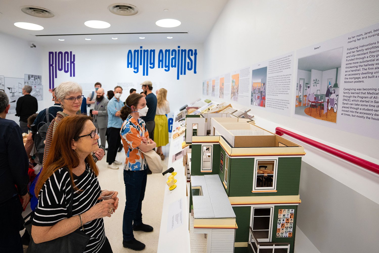 A crowd looks at architecture models and drawings in a gallery space.