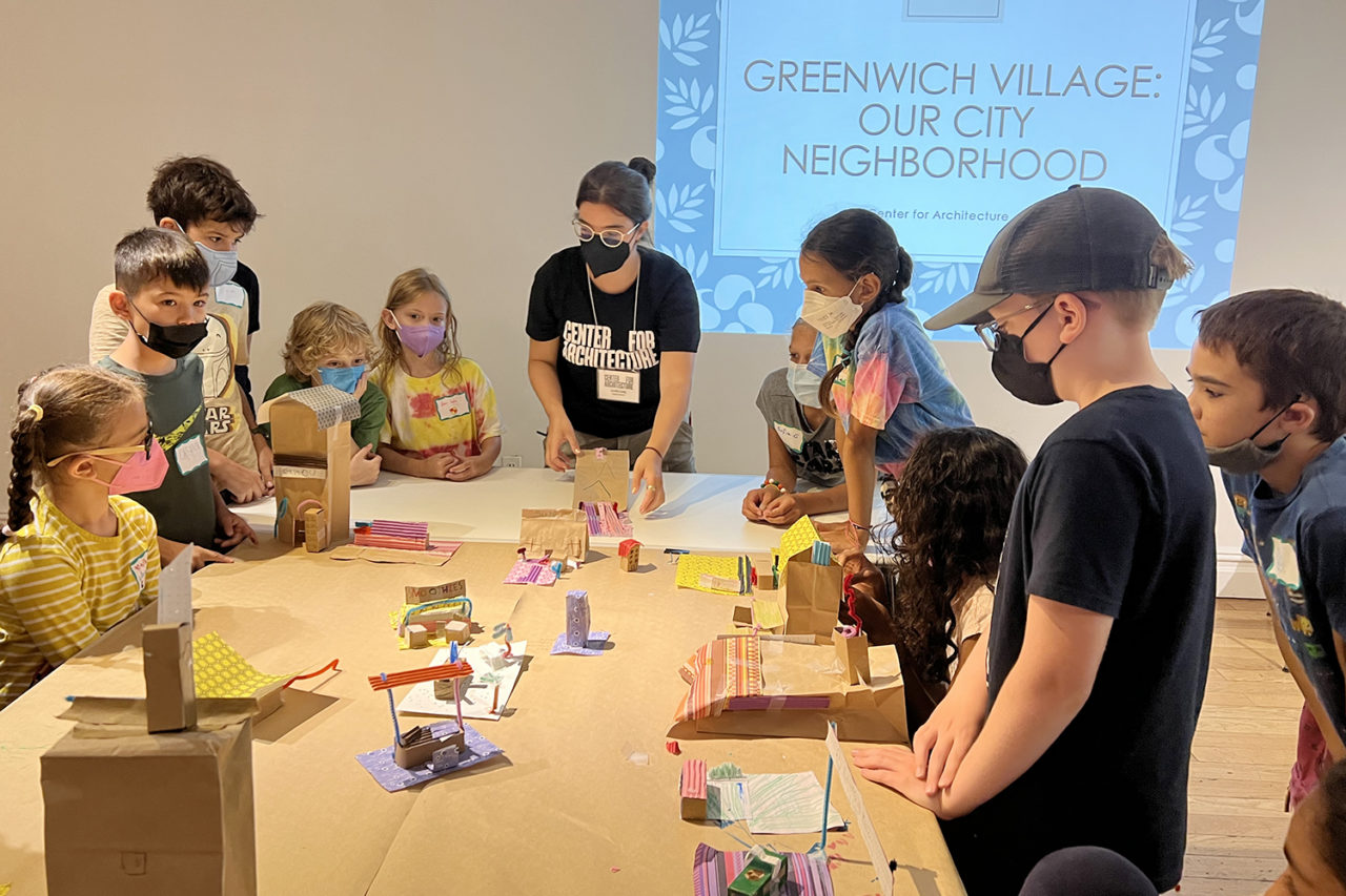 Woman surrounded by students demonstrates model-building activity.