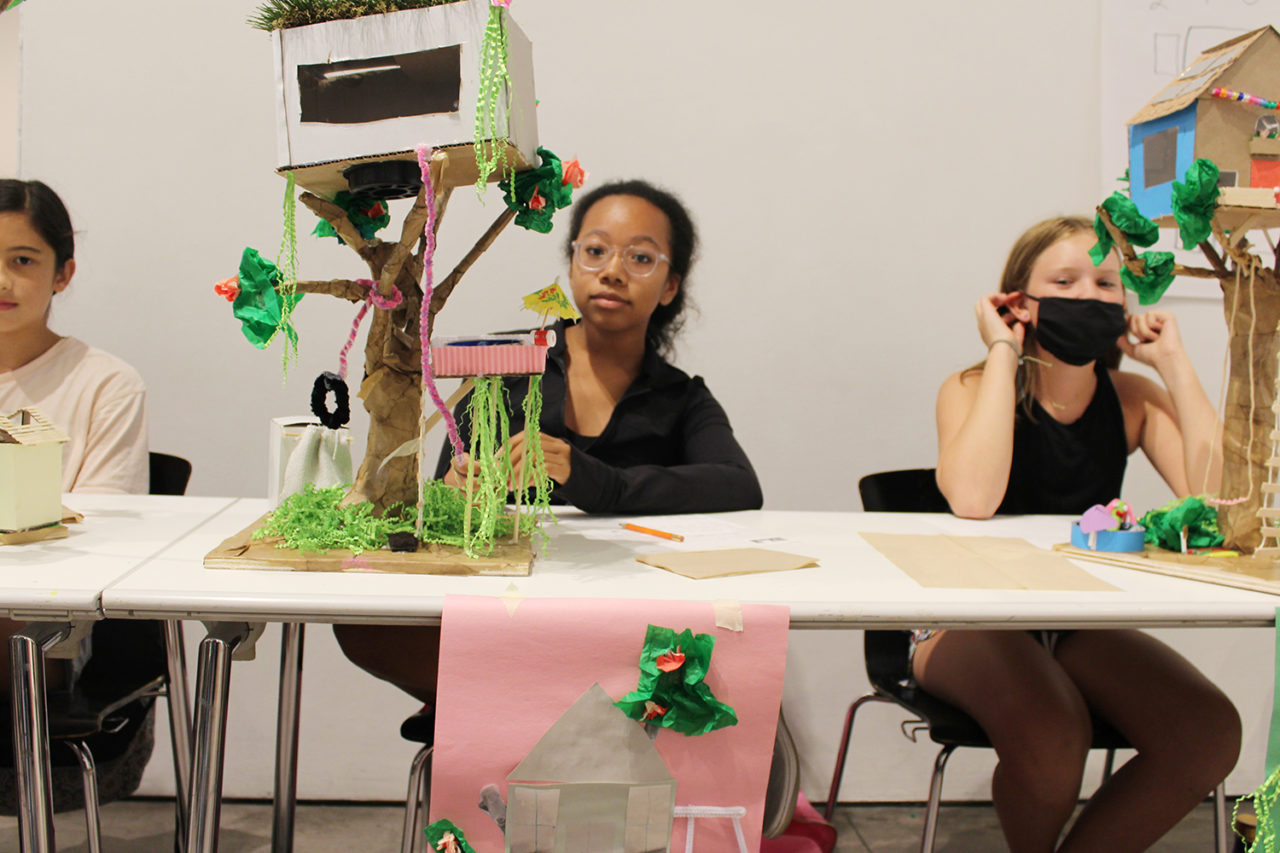 Three students stand behind models of treehouses.