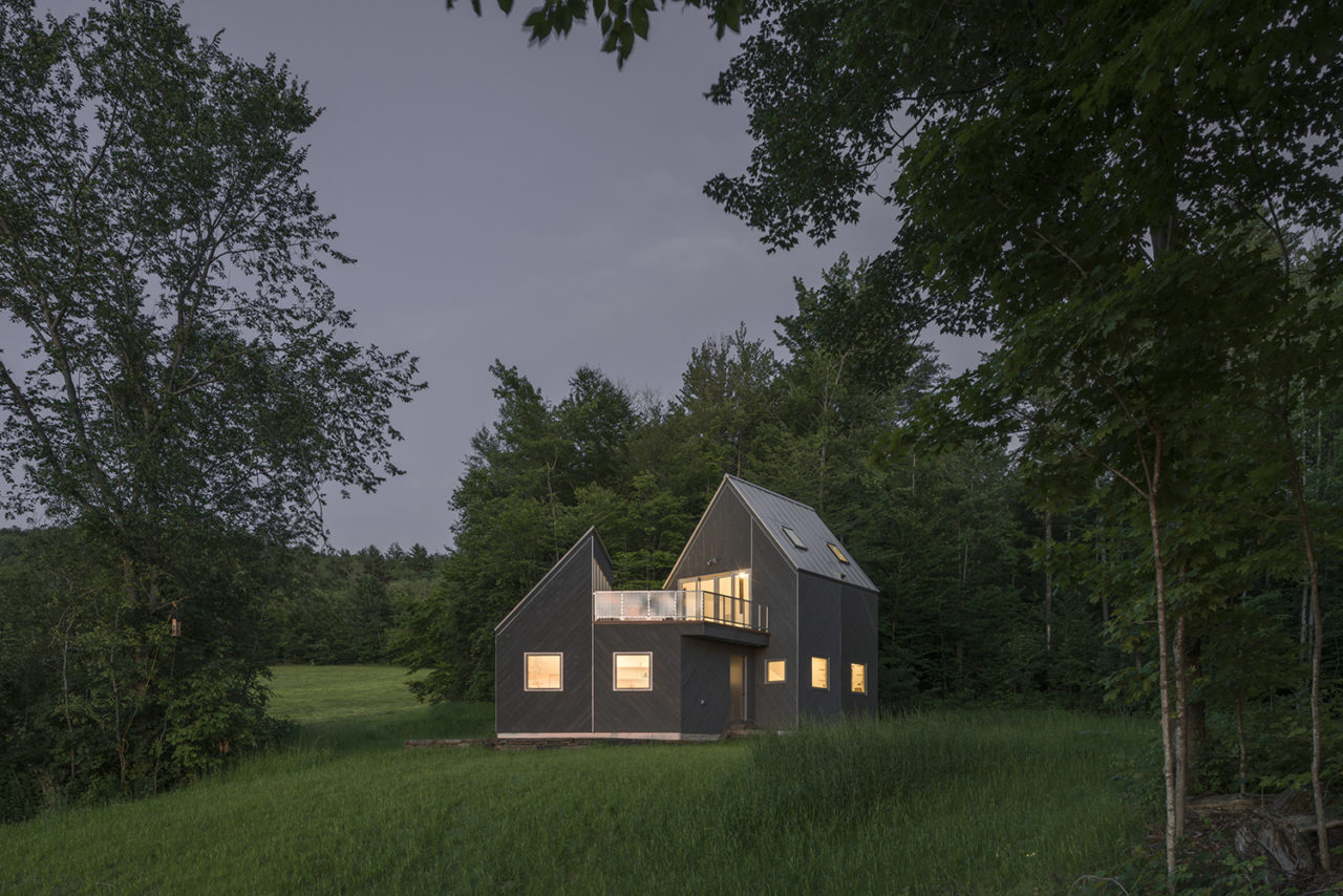 A grey-clad cabin set amidst greenery, lit from within at dusk
