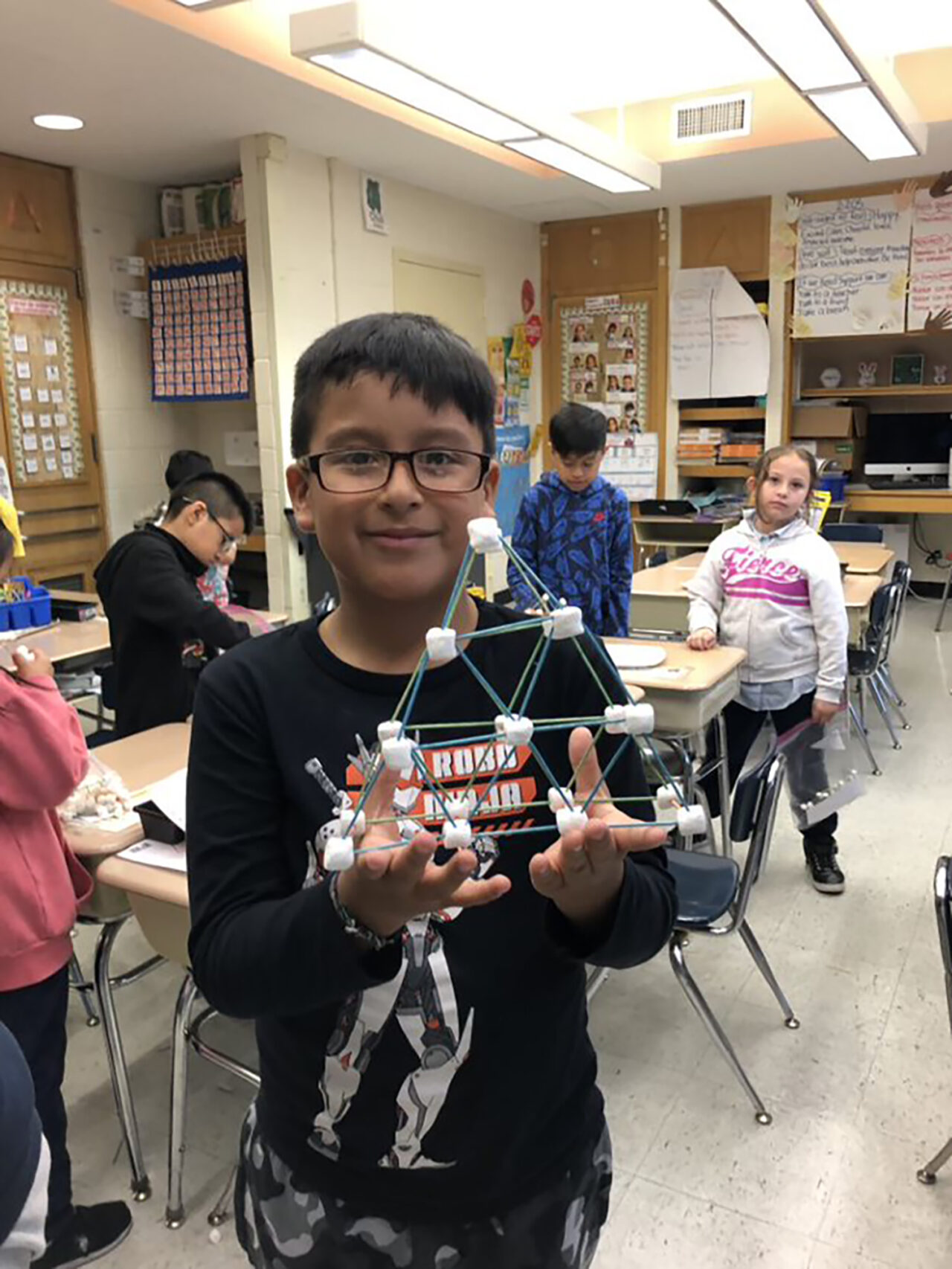 A young boy holds up a pyramid-structured truss bridge made with sticks and marshmallows.