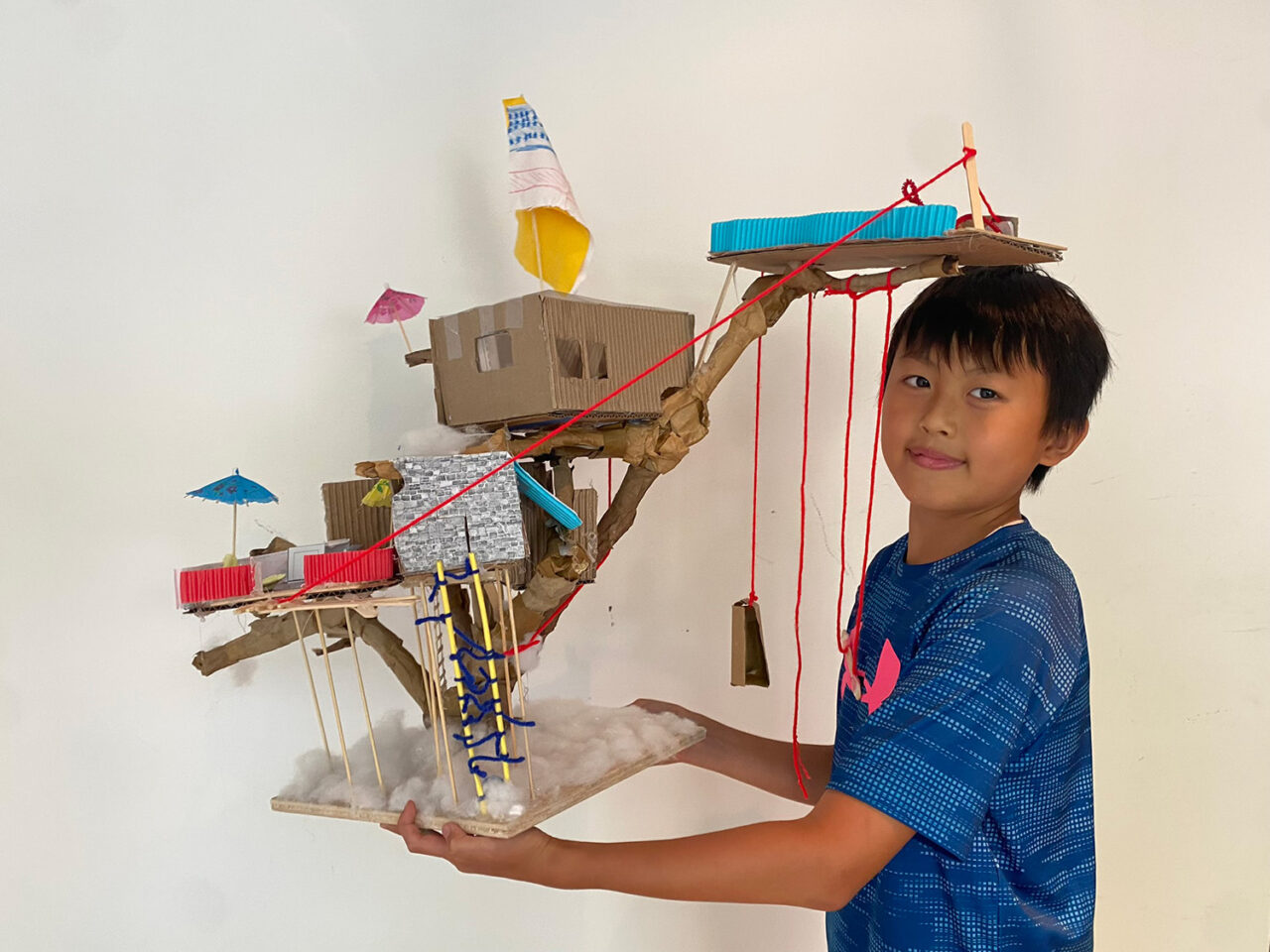 Boy wearing a blue shirt poses with his model tree house, a three-platformed cardboard-wood structure with yarn hanging from its levels.
