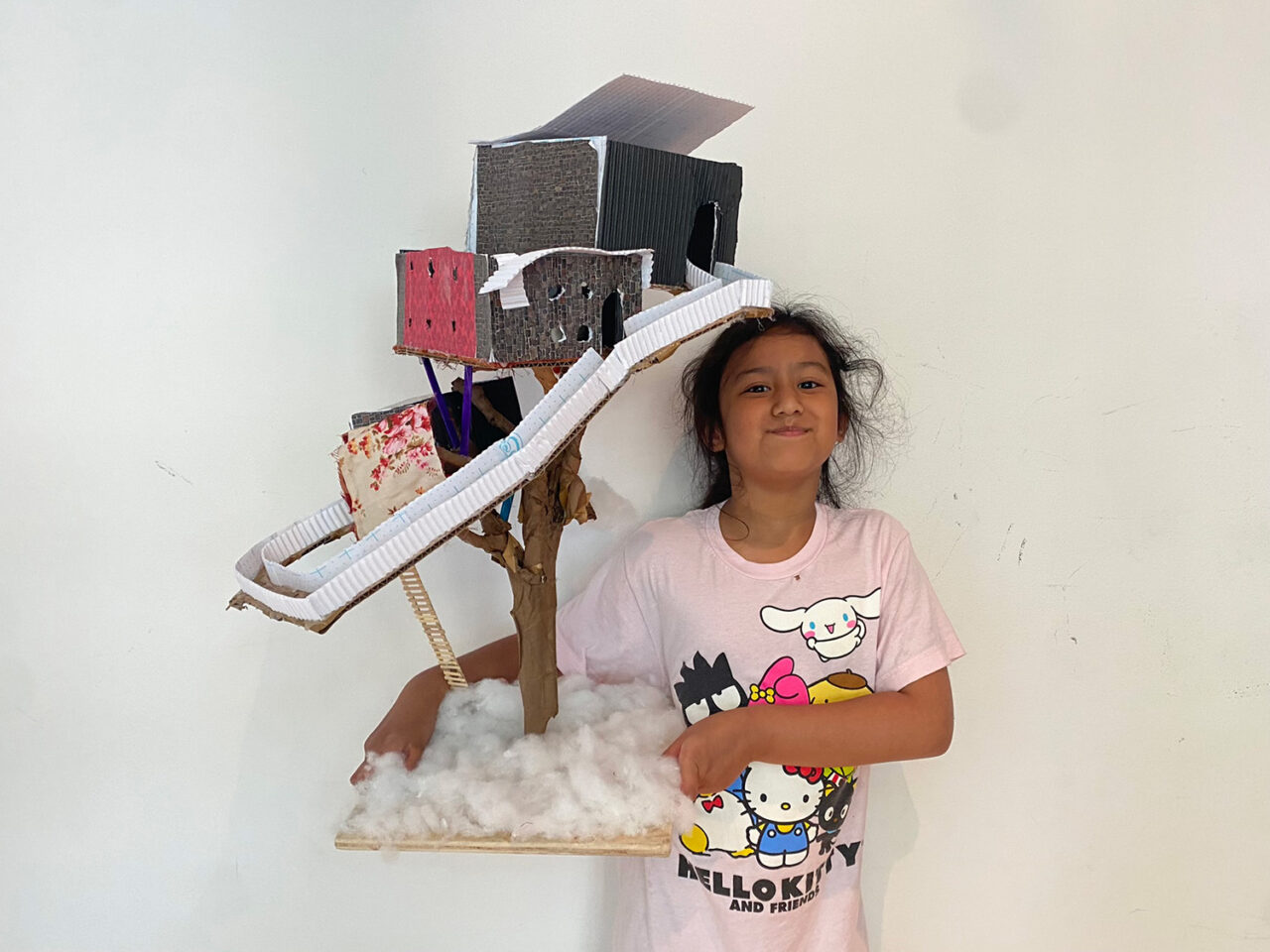 Girl in a pink shirt poses with a two story model tree house. Cotton balls cover the wooden base.