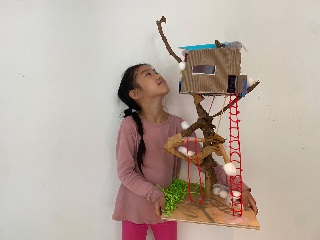 Girl in a pink long sleeve shirt gazes up at her tree house structure, built on a tall wooden branch with a red pipe cleaner ladder leading to the base.