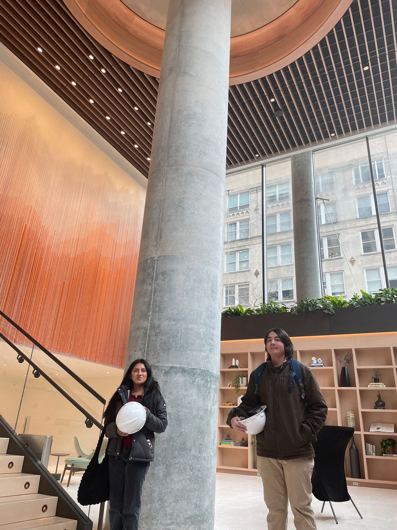 Two students carrying hard hats and standing inside a tall ceilinged interior with a concrete column