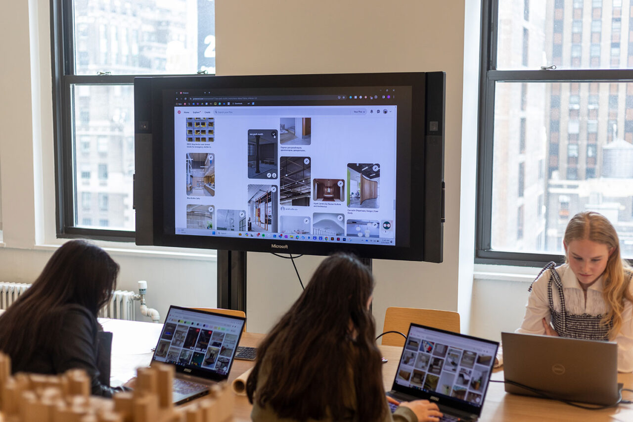 Students with laptops sit at a conference table and reference a larger screen with various images on it
