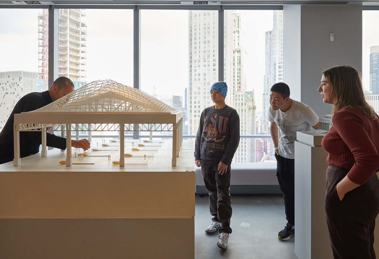 Students gather around a large architectural model with instruction from an adult