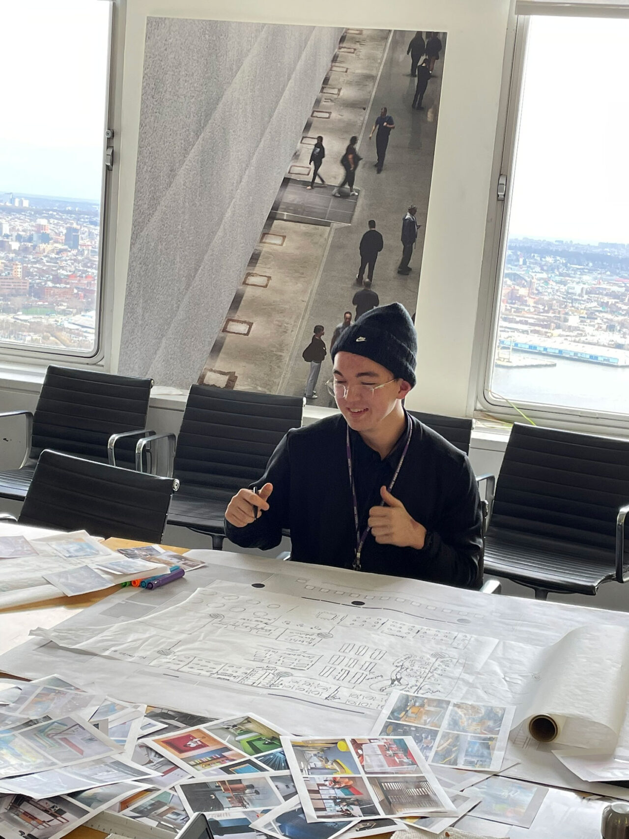 A student sits at a conference table surrounded by architectural drawings and smiles