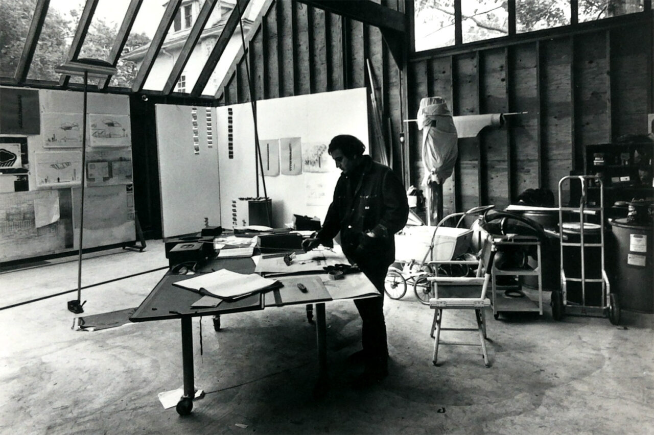 A black and white photo of a large sculpture studio with large glass windows above. A man stands at the desk, writing something.