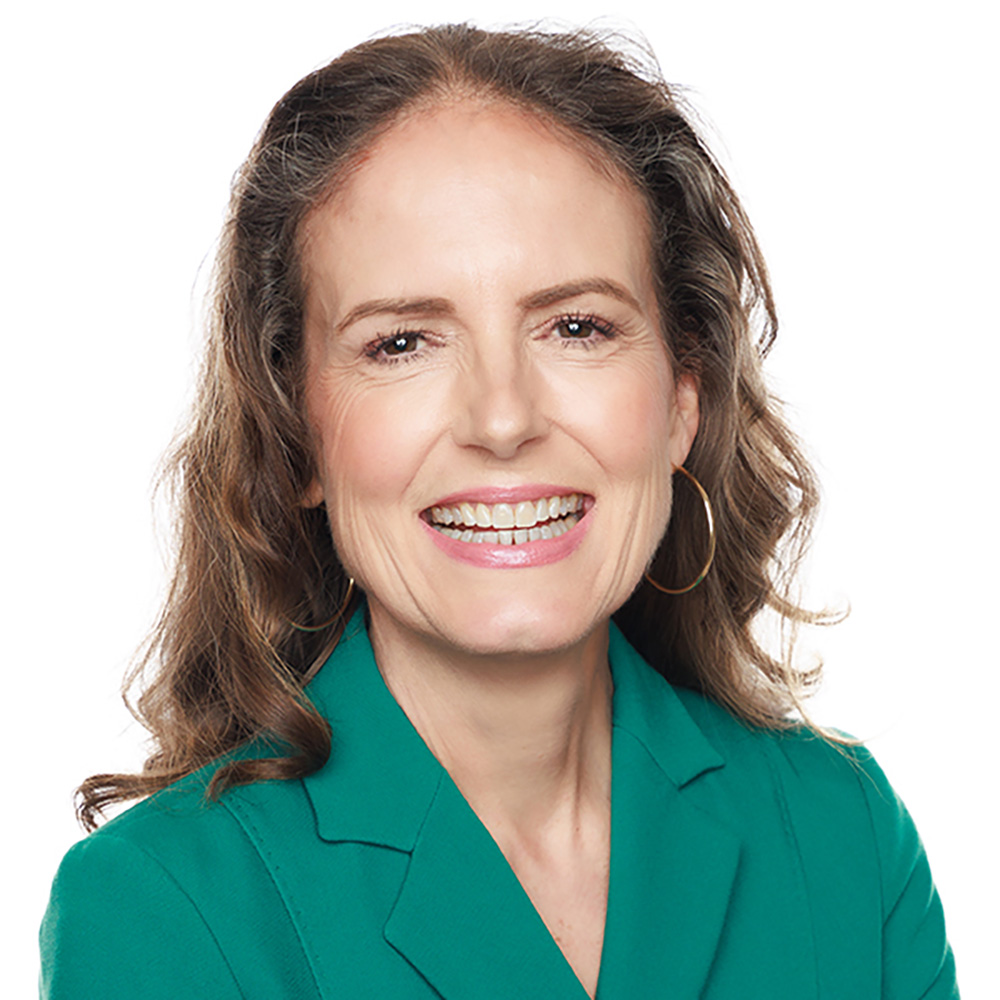 Headshot of a white woman with wavy brown hair wearing a green blazer.