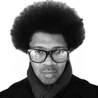 Black and white headshot of a Black man wearing dark framed glasses and a scarf.
