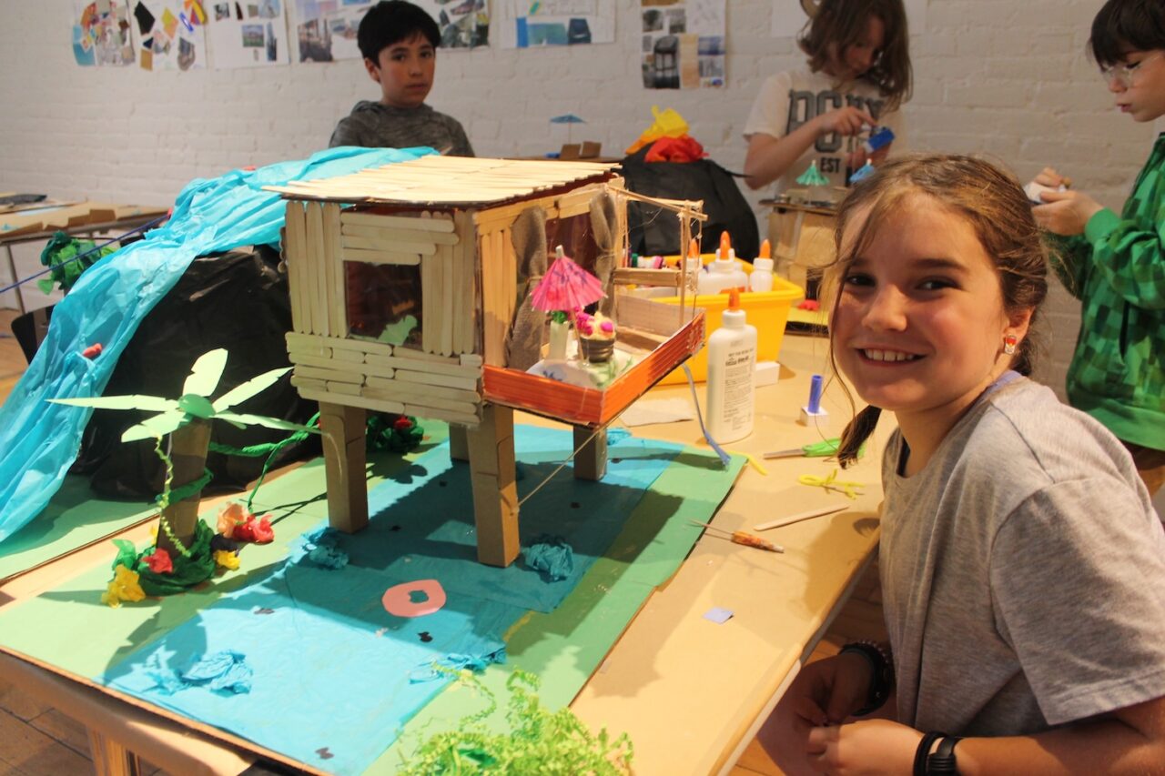 One elementary school student smiling next to their model of a wooden house over water.