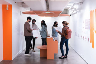 Installation view with five people standing and reading exhibition content behind an orange seat with an orange trellis installed above them