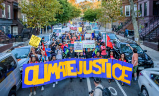 A group of protestors or marchers standing in a residential looking street holding a large banner that reads "Climate Justice"