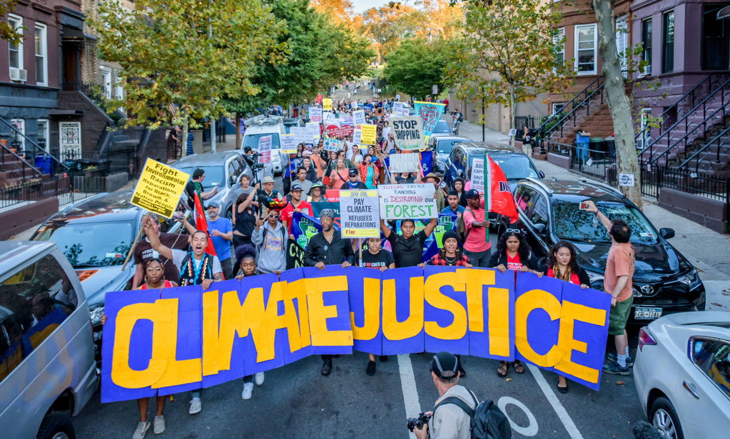 A group of protestors or marchers standing in a residential looking street holding a large banner that reads "Climate Justice"